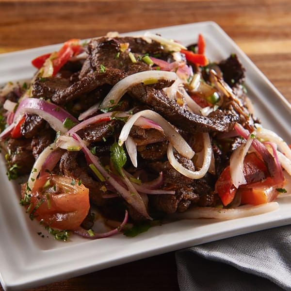 A close-up of a dish with sliced beef, red onions, tomatoes, and herbs on a white plate.