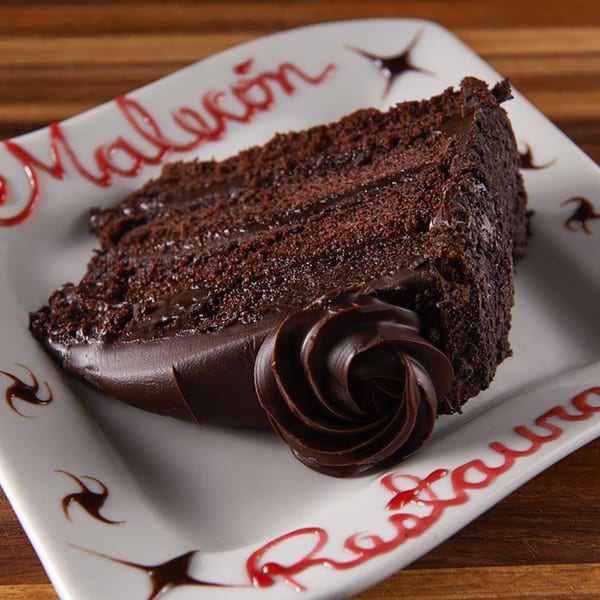 A slice of rich chocolate cake with chocolate frosting and a chocolate swirl on a white plate.