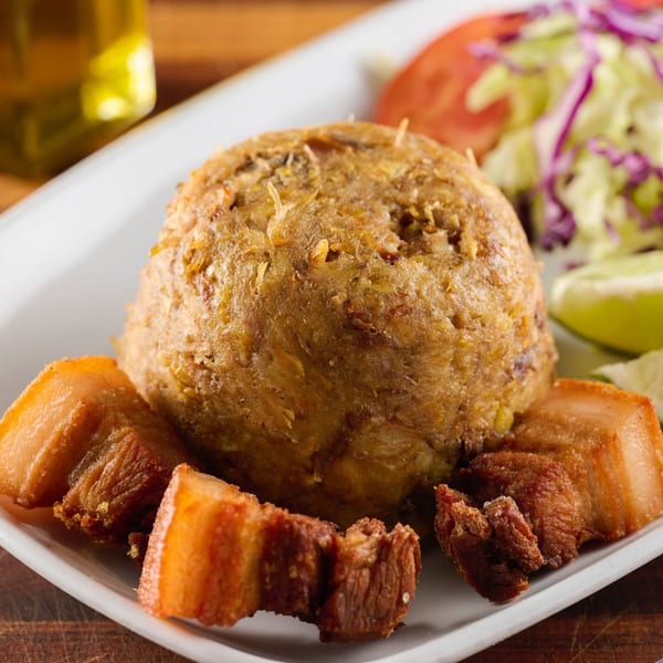 Mofongo with fried pork chunks, a side salad, and lime wedges on a white plate.