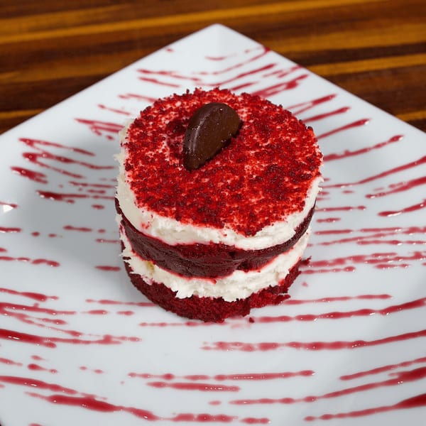Red velvet cake with white frosting layers, topped with red sprinkles and a chocolate garnish.