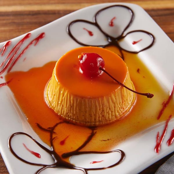 Golden flan dessert with caramel sauce and a maraschino cherry on a white plate with chocolate and raspberry drizzle.