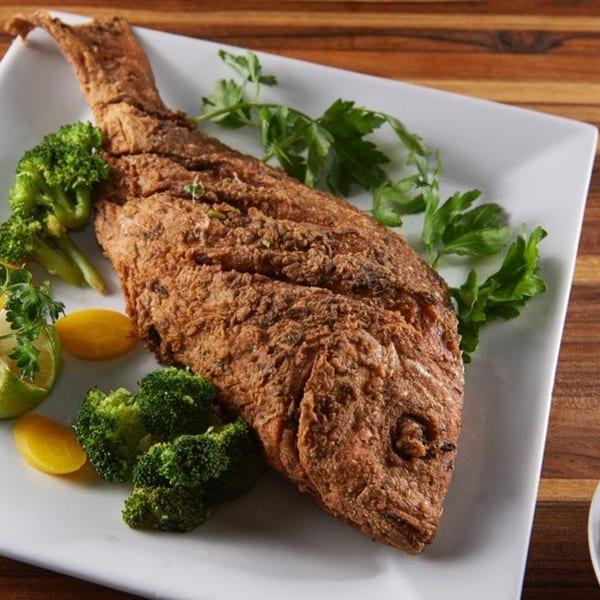 Whole fried fish with crispy skin, garnished with broccoli, lime, and parsley on a white plate.