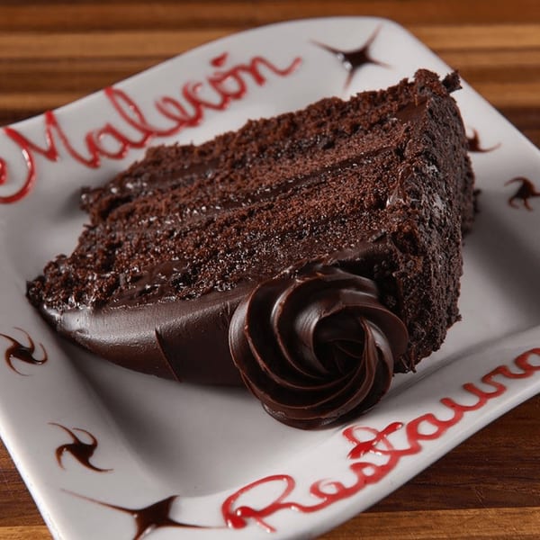 A slice of rich chocolate cake with frosting and a chocolate rosette on a white plate.